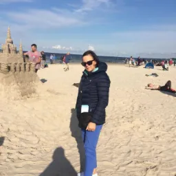 Kobieta w okularach przeciwsłonecznych i kurtce pozuje na plaży obok budowli z piasku w słoneczny dzień nad morzem.