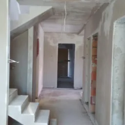 Wnętrze domu w trakcie tynkowania: świeżo otynkowane ściany i schody, widoczne instalacje elektryczne, surowy stan wykończenia, perspektywa korytarza.