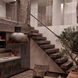 ArtCade Studio - Loftowe wnętrze z masywnymi, surowymi drewnianymi belkami i schodami, szklaną balustradą, industrialną lampą oraz aneksem kuchennym z metalicznymi frontami. Widoczna sofa i roślina w donicy.