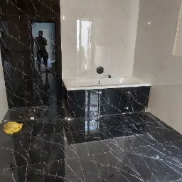 Wykończona łazienka z czarno-białymi płytkami imitującymi marmur, z widoczną wanną i niedokończonym oświetleniem sufitowym, odbicie osoby w błyszczącej powierzchni płytek.