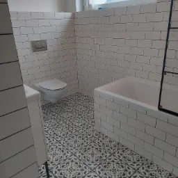 Biała łazienka z czarno-białą wzorzystą podłogą, białe płytki na ścianach ułożone w cegiełkę, wisząca toaleta, wanna i umywalka, widok przez okno na dachy budynków.