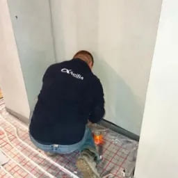 Pracownik w ciemnej bluzie z logo firmy Agilia instaluje ogrzewanie podłogowe na folii refleksyjnej z czerwoną siatką w rogu pomieszczenia z surowymi ścianami.