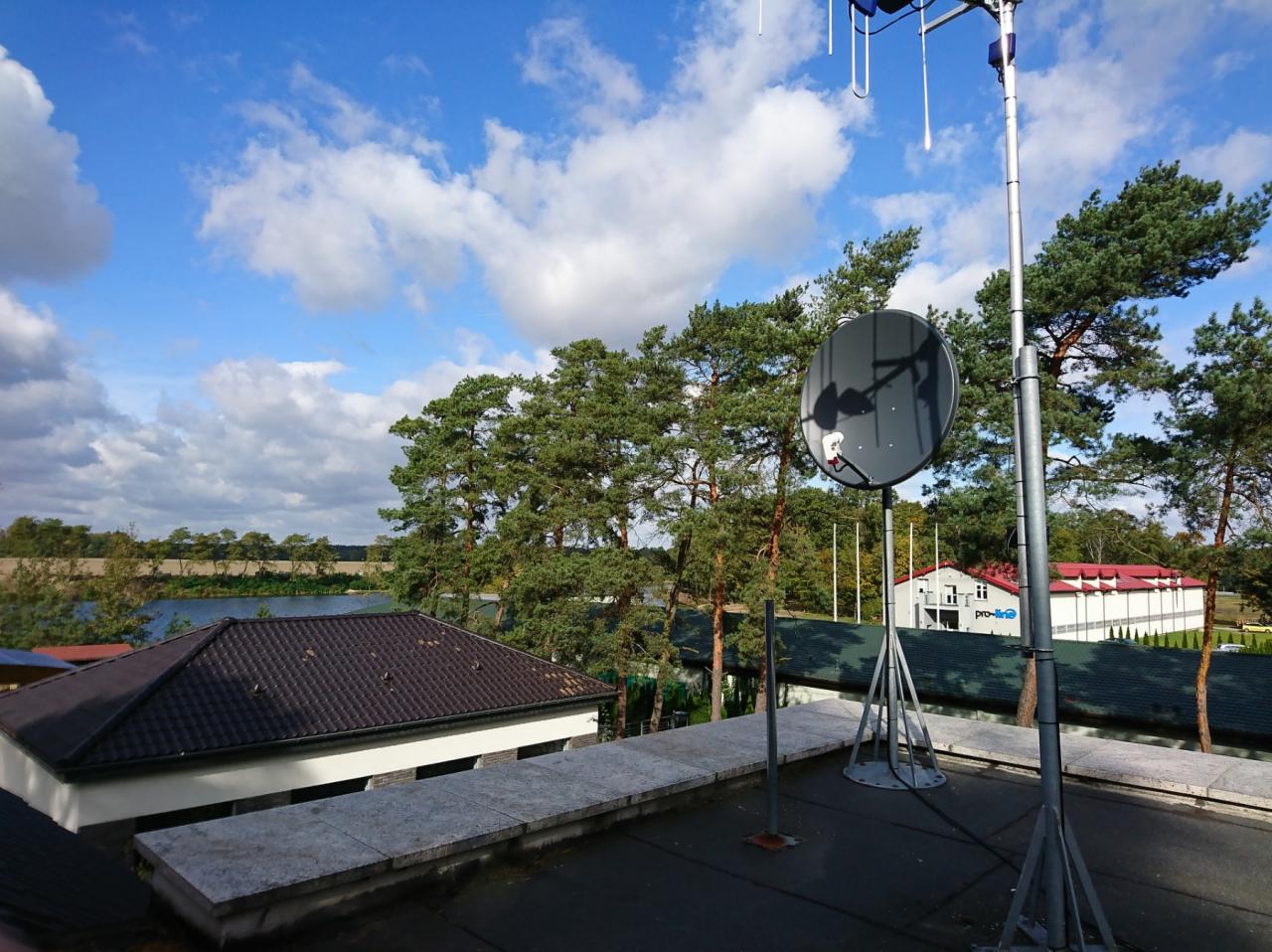 Antena satelitarna zamontowana na dachu budynku, widok z góry na otaczający krajobraz z drzewami i jeziorem w tle, błękitne niebo z białymi chmurami.