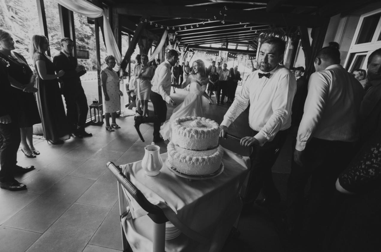 Czarno-białe zdjęcie z wesela: Para młoda tańczy w tle, a na pierwszym planie widać dwupoziomowy tort na wózku, z dzbankiem obok, na tle gości weselnych i dekoracji.