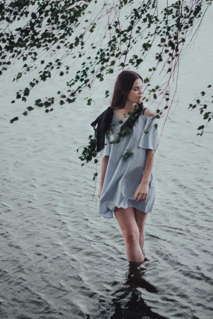Młoda kobieta w pasiastej sukience stoi w wodzie, z gałązkami drzewa zwisającymi nad nią, tworząc naturalną ramę wokół postaci.