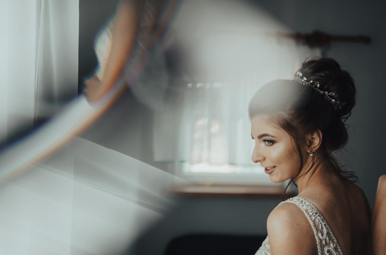 Panna młoda z upiętymi włosami i ozdobną opaską, spogląda w bok, widoczna przez rozmyte odbicie w lustrze, w tle okno z drewnianą ramą.