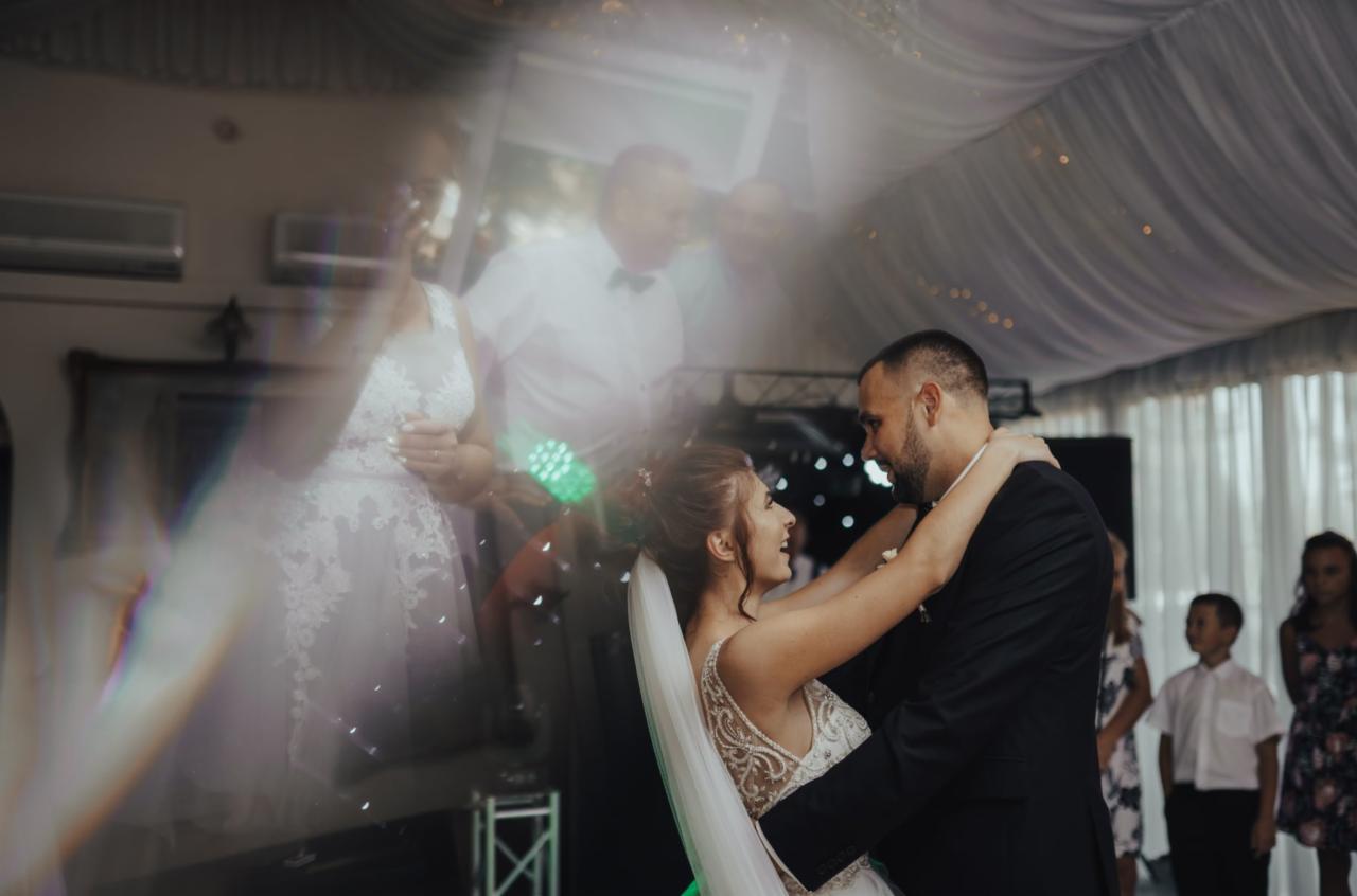 Para młoda tańczy w objęciach na tle dekoracji sali weselnej i obserwujących gości.