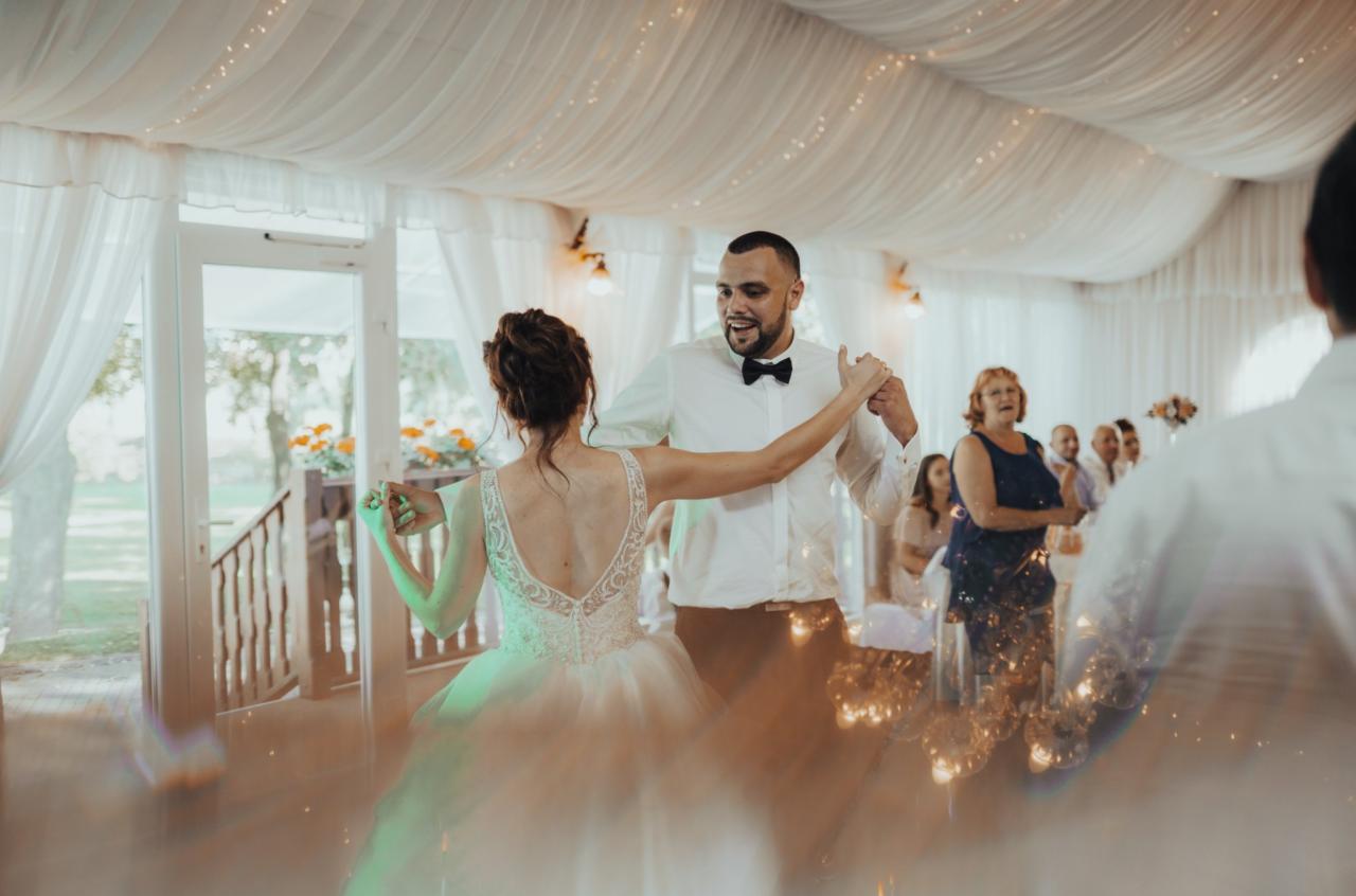 Para młoda tańczy taniec weselny w udekorowanej sali. Panna młoda w sukni z koronkowymi plecami, pan młody w białej koszuli z muszką. W tle goście weselni i dekoracje świetlne.