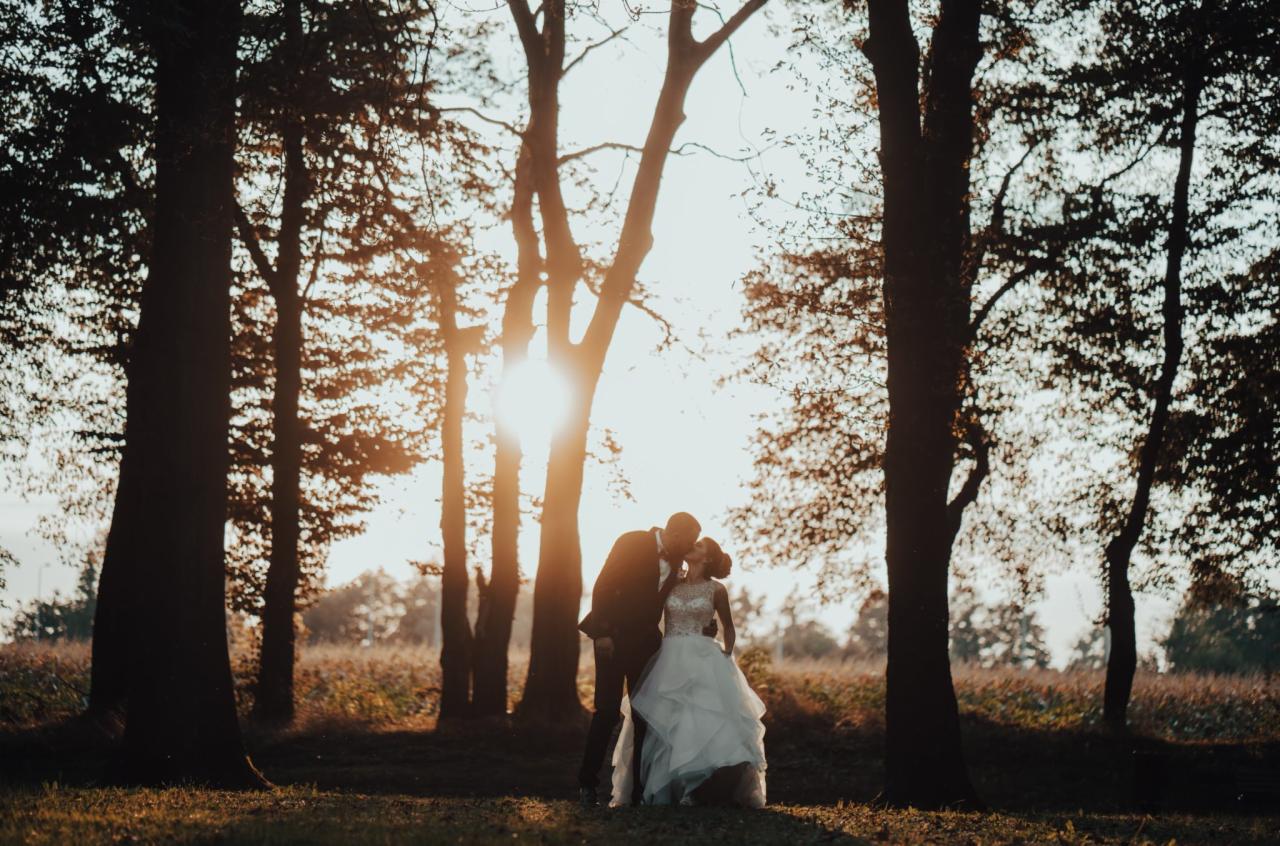 Para młoda całująca się w lesie podczas złotej godziny, suknia ślubna z koronkową górą i spódnicą z falban, ciemny garnitur, drzewa w tle, ciepłe światło słoneczne.