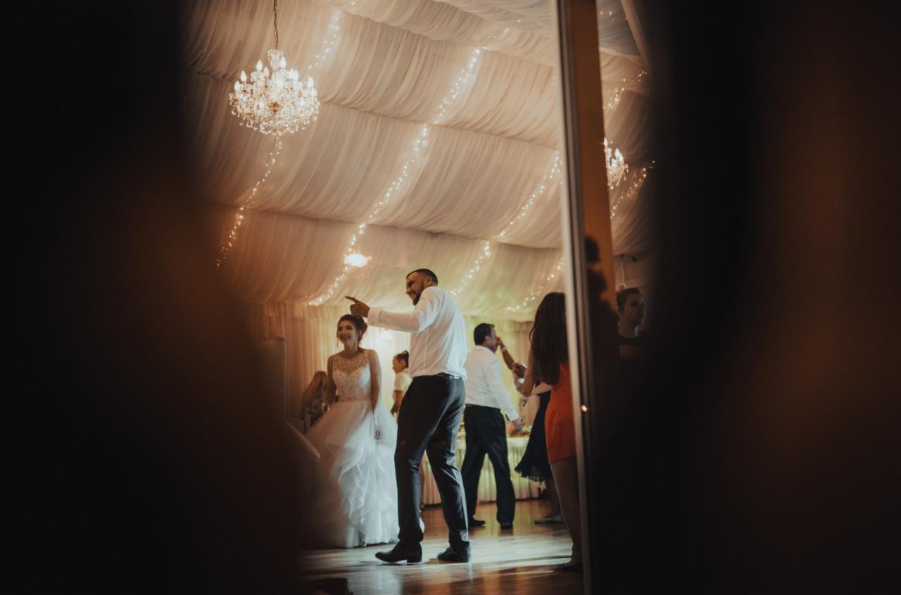 Para młoda na parkiecie, mężczyzna w białej koszuli i ciemnych spodniach wskazuje palcem, w tle dekoracja sali z białych materiałów i kryształowy żyrandol.