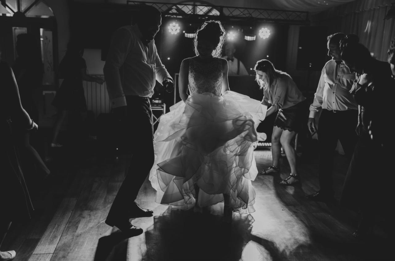 Czarno-białe zdjęcie tańczącej panny młodej w sukni z falbanami, otoczonej przez gości weselnych na parkiecie oświetlonym reflektorami.