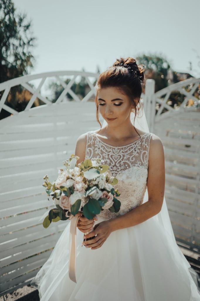 Panna młoda w białej sukni z koronkową górą, trzyma bukiet różowych i białych róż z satynową wstążką, na tle białego, ażurowego płotu.