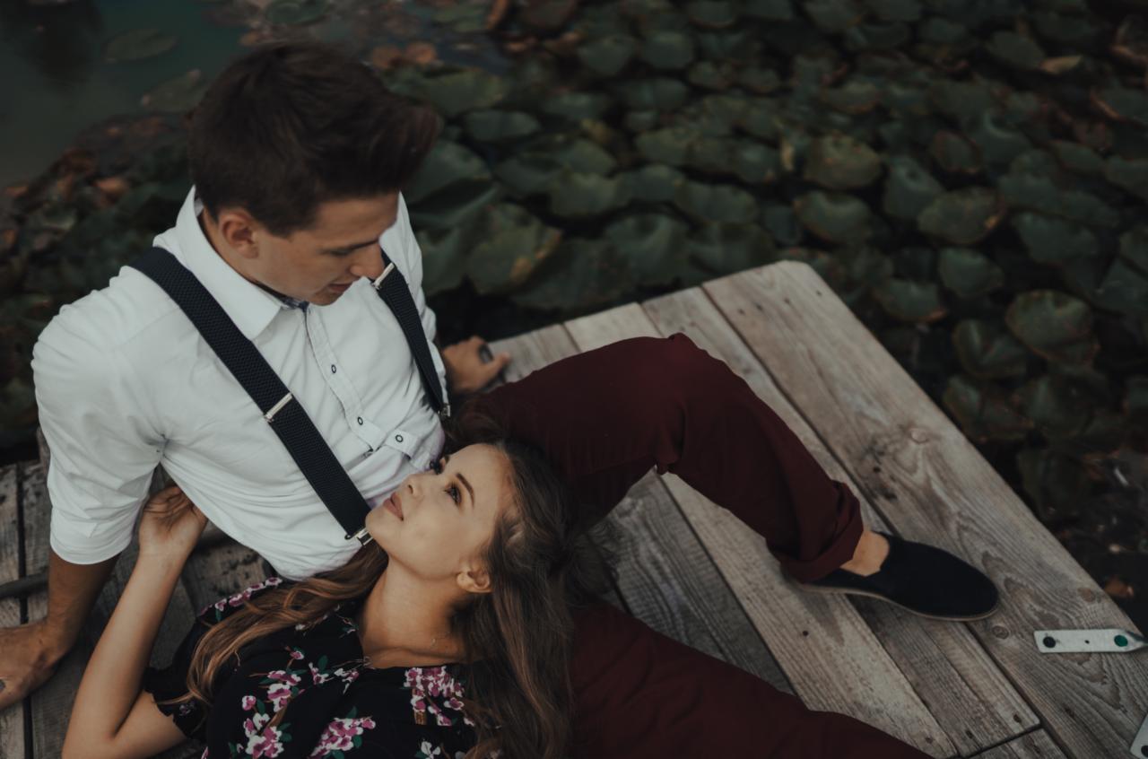 Zakochana para leży na drewnianym pomoście nad wodą porośniętą liliami wodnymi. Mężczyzna w białej koszuli i szelkach patrzy na leżącą na jego nogach kobietę w kwiecistej sukience.