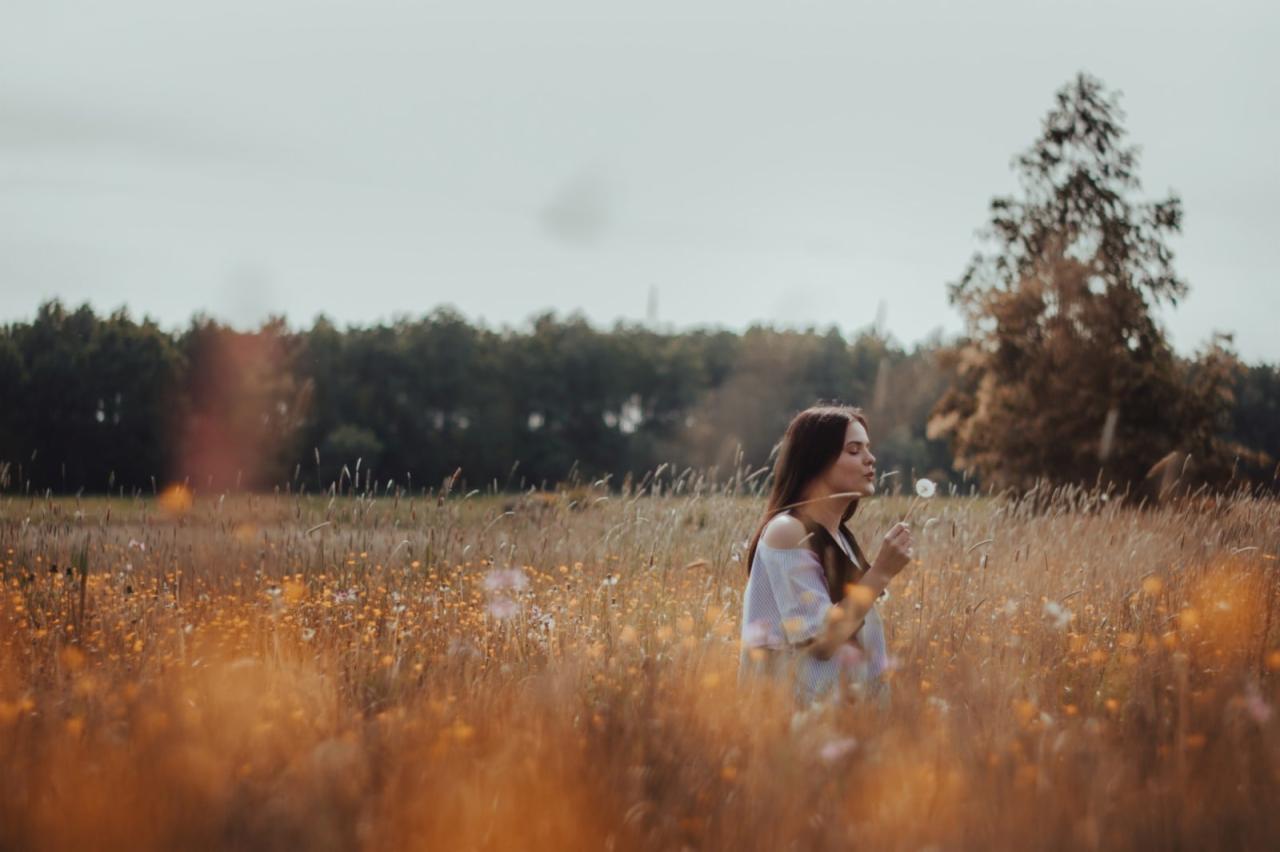 Młoda kobieta z długimi brązowymi włosami, ubrana w bluzkę z odkrytymi ramionami, dmucha na dmuchawca na łące pełnej wysokiej trawy i drobnych żółtych kwiatów, z lasem i pojedynczym drzewem w tle.