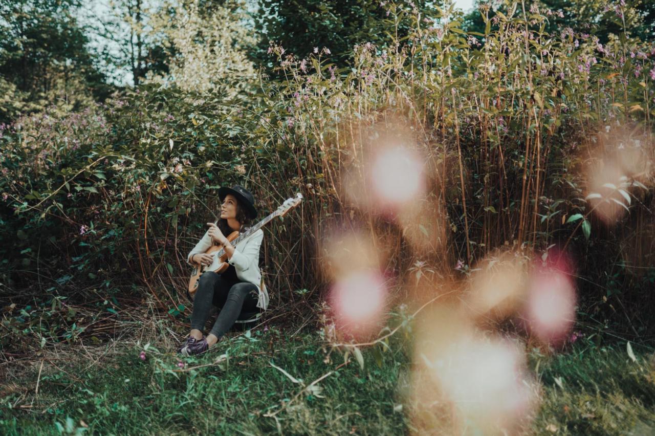 Kobieta w kapeluszu siedzi na trawie, trzymając gitarę basową, w tle zarośla i drzewa, z efektem bokeh na pierwszym planie.
