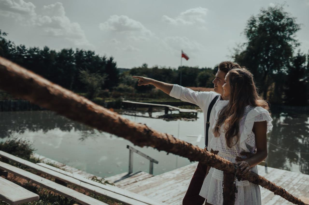 Para na drewnianym pomoście, mężczyzna wskazuje palcem w dal, kobieta trzyma się drewnianej barierki, w tle woda i drzewa, flaga w oddali.