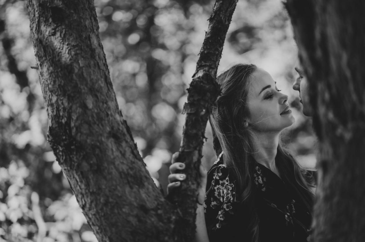 Czarno-białe zdjęcie pary zakochanych, kobieta opiera się o drzewo, mężczyzna stoi naprzeciwko niej, zbliżeni twarzami, delikatny uśmiech kobiety, naturalne otoczenie drzew.
