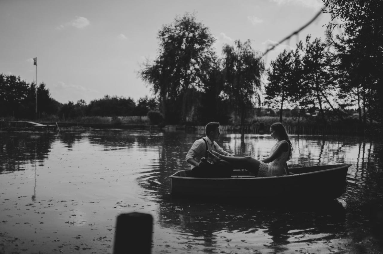 Czarno-białe zdjęcie pary w łódce na jeziorze; mężczyzna w koszuli i szelkach trzyma wiosło, kobieta w koronkowej sukience siedzi obok niego; w tle drzewa i flaga.