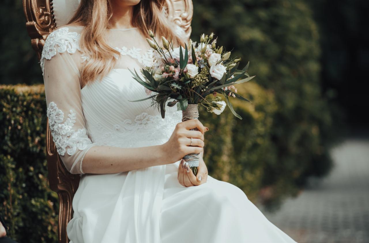 Panna młoda w białej sukni z koronkowymi rękawami, siedząca na złotym krześle i trzymająca bukiet ślubny z białych i różowych kwiatów, z zielonym tłem.