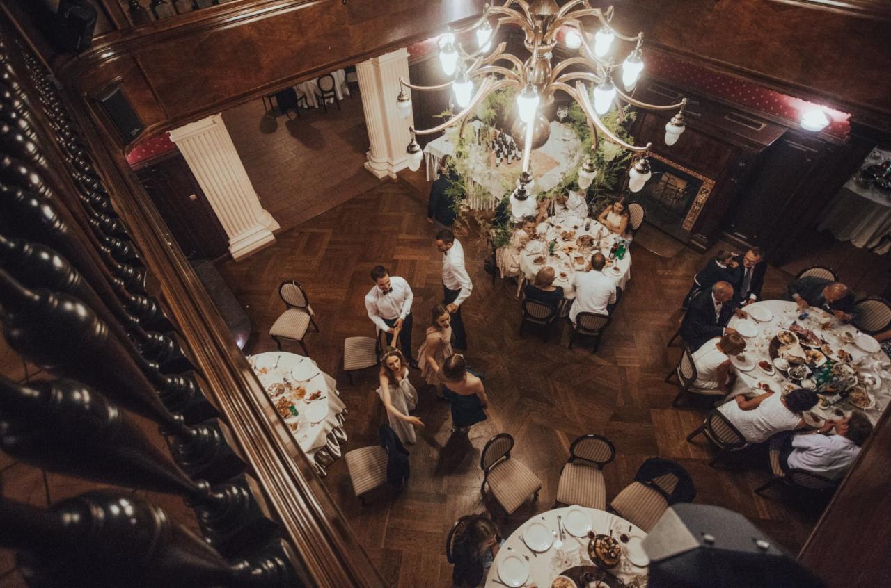 Widok z góry na salę balową z gośćmi siedzącymi przy okrągłych stołach, parkiet, dekoracje kwiatowe i żyrandol, widziane przez balustradę.