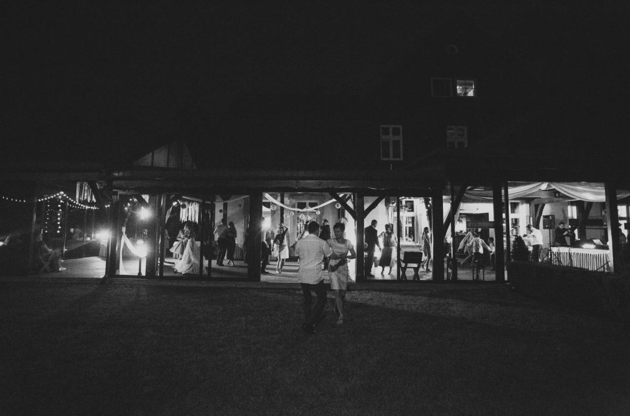Czarno-białe zdjęcie pary tańczącej na trawie przed budynkiem z oświetlonymi oknami, wewnątrz widoczni goście i dekoracje, nocne ujęcie z niskim oświetleniem.