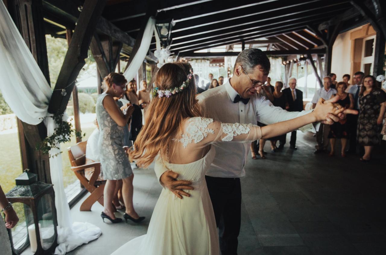 Para tańczy podczas uroczystości, kobieta w białej sukni z koronkowymi rękawami i wiankiem na głowie, mężczyzna w białej koszuli z muchą, w tle goście i dekoracje.