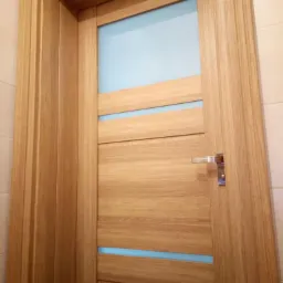 Drewniane drzwi wewnętrzne z przeszkleniami w kolorze błękitnym, widoczna klamka i ościeżnica, na dole logo firmy.