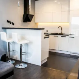 Nowoczesna, minimalistyczna kuchnia z białymi, lakierowanymi szafkami, czarnym blatem i barkiem z dwoma białymi hokerami, widoczny fragment szarej sofy