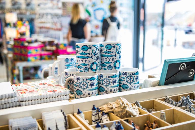Półka z pamiątkami w sklepie: kubki z motywami morskimi (ryby, kotwice, koła sterowe), notatniki z nadrukami, miniaturowe latarnie morskie i inne drobne upominki.