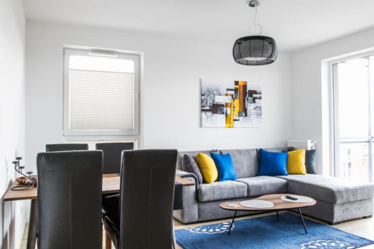 Jasne wnętrze mieszkania z szarą sofą narożną, niebieskim dywanem i drewnianym stołem jadalnym z ciemnymi krzesłami.