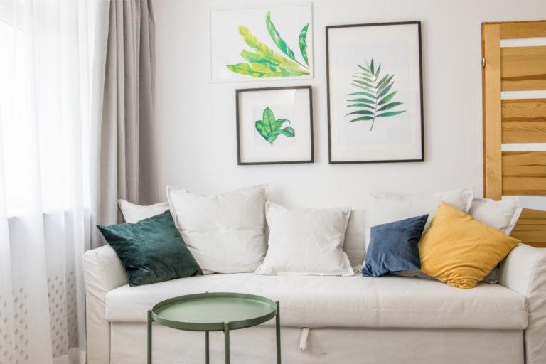 Jasny salon z białym narożnikiem, kolorowymi poduszkami, zielonym stolikiem kawowym i botanicznymi grafikami na ścianie.