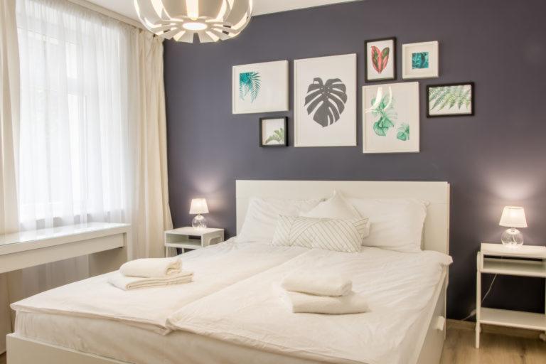 Jasna sypialnia z podwójnym łóżkiem, białymi meblami i dekoracjami ściennymi z motywami roślinnymi.