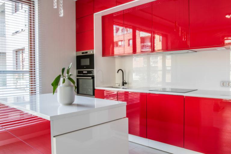 Nowoczesna kuchnia z czerwonymi, lakierowanymi szafkami, białym blatem i wyspą kuchenną z rośliną w białym wazonie.