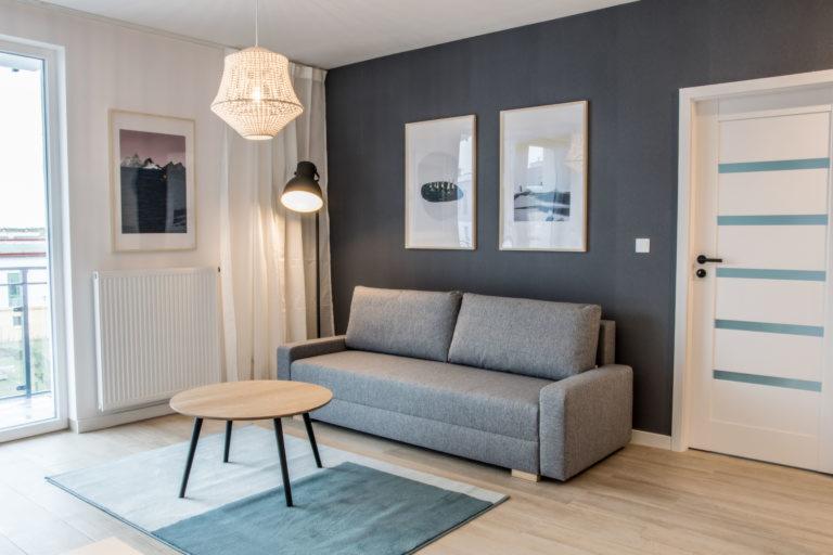 Jasne, nowocześnie urządzone wnętrze pokoju dziennego z szarą sofą, okrągłym drewnianym stolikiem na dywanie w odcieniach błękitu i bieli, wiszącą lampą z plecionki oraz obrazami na ciemnoszarej...