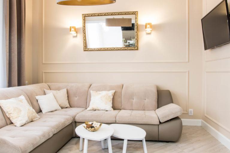 Jasny salon z narożną sofą, dwoma okrągłymi stolikami, lustrem w złotej ramie i telewizorem na ścianie.