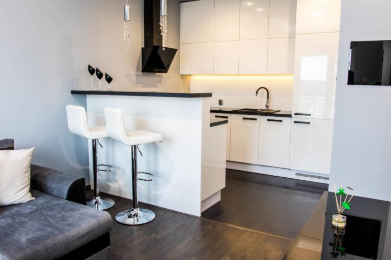 Nowoczesna, minimalistyczna kuchnia z białymi, lakierowanymi szafkami, czarnym blatem i barkiem z dwoma białymi hokerami, widoczny fragment szarej sofy