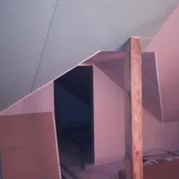 Poddasze w trakcie prac wykończeniowych, widoczne płyty gipsowo-kartonowe na skośnych ścianach i suficie, drewniany słup podtrzymujący konstrukcję, oraz przewody i materiały budowlane na podłodze.