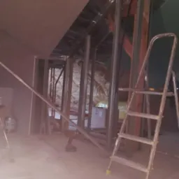 Wnętrze poddasza w trakcie adaptacji: widoczne metalowe profile konstrukcyjne ścian działowych, częściowo zamontowane płyty kartonowo-gipsowe na skosach, drabina aluminiowa i narzędzia budowlane...