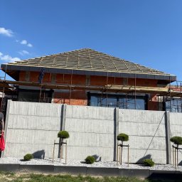 Usługi Dekarsko-Ogólnobudowlane - Dom w trakcie budowy z widocznym szkieletem dachu, rusztowaniami i betonowym ogrodzeniem. Niebo błękitne, słoneczny dzień. Małe drzewka ozdobne przed ogrodzeniem.