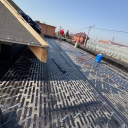 Usługi Dekarsko-Ogólnobudowlane - Czarny dach pokryty papą z widocznym wzorem, na którym stoją butle gazowe i narzędzia. W tle domy i szklarnia, a w oddali pracownik w czerwonej koszulce.