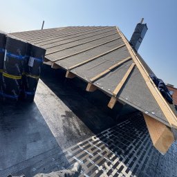 Usługi Dekarsko-Ogólnobudowlane - Dach w trakcie krycia papą na deskowaniu, widoczne bale drewniane, komin zabezpieczony folią, z boku rolki papy. Ujęcie z dołu, w kadrze noga dekarza w kombinezonie.