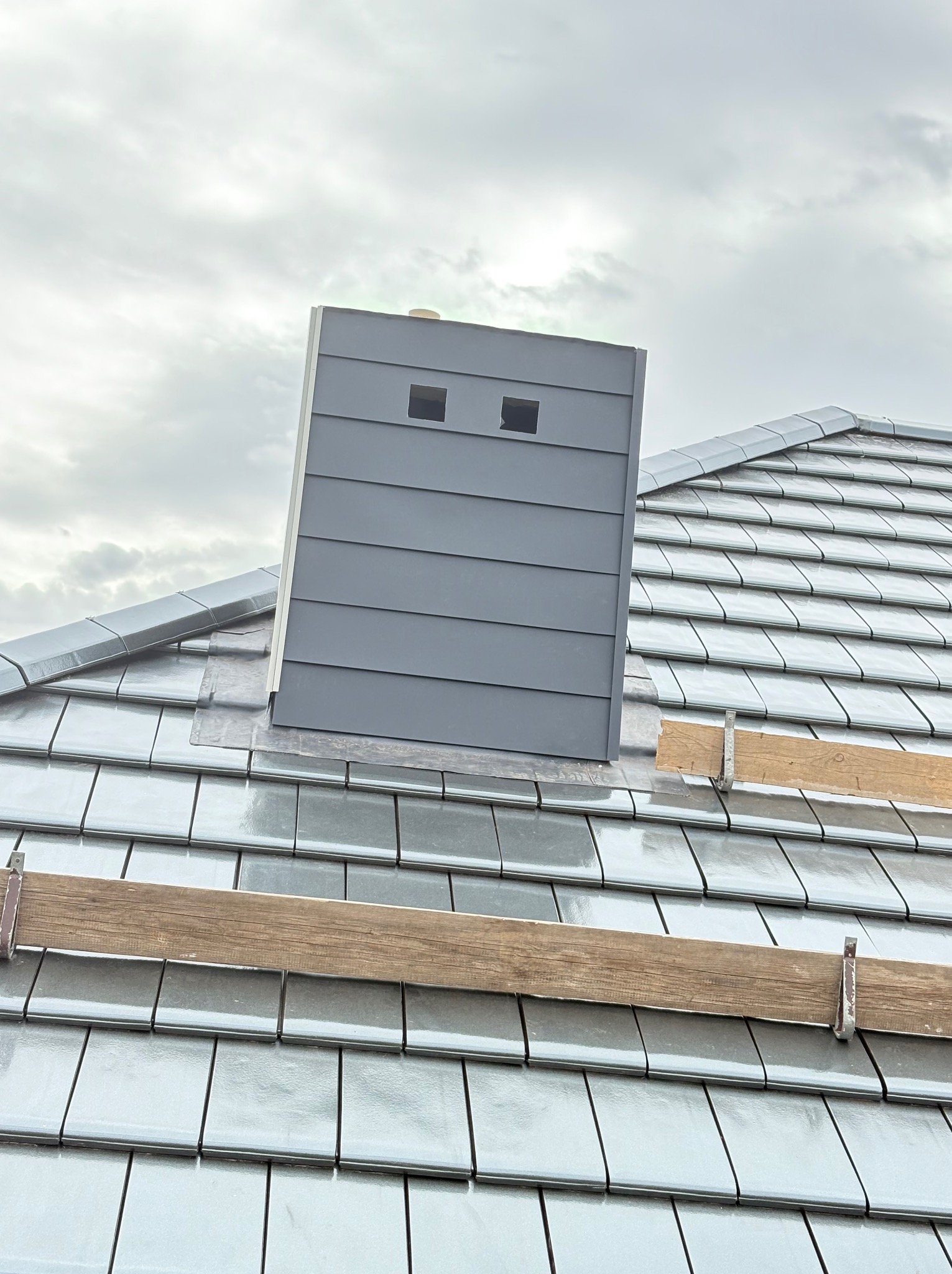 Szary komin wentylacyjny na dachu z błyszczącej, szarej dachówki ceramicznej. Widoczne drewniane elementy rusztowania i pochmurne niebo.