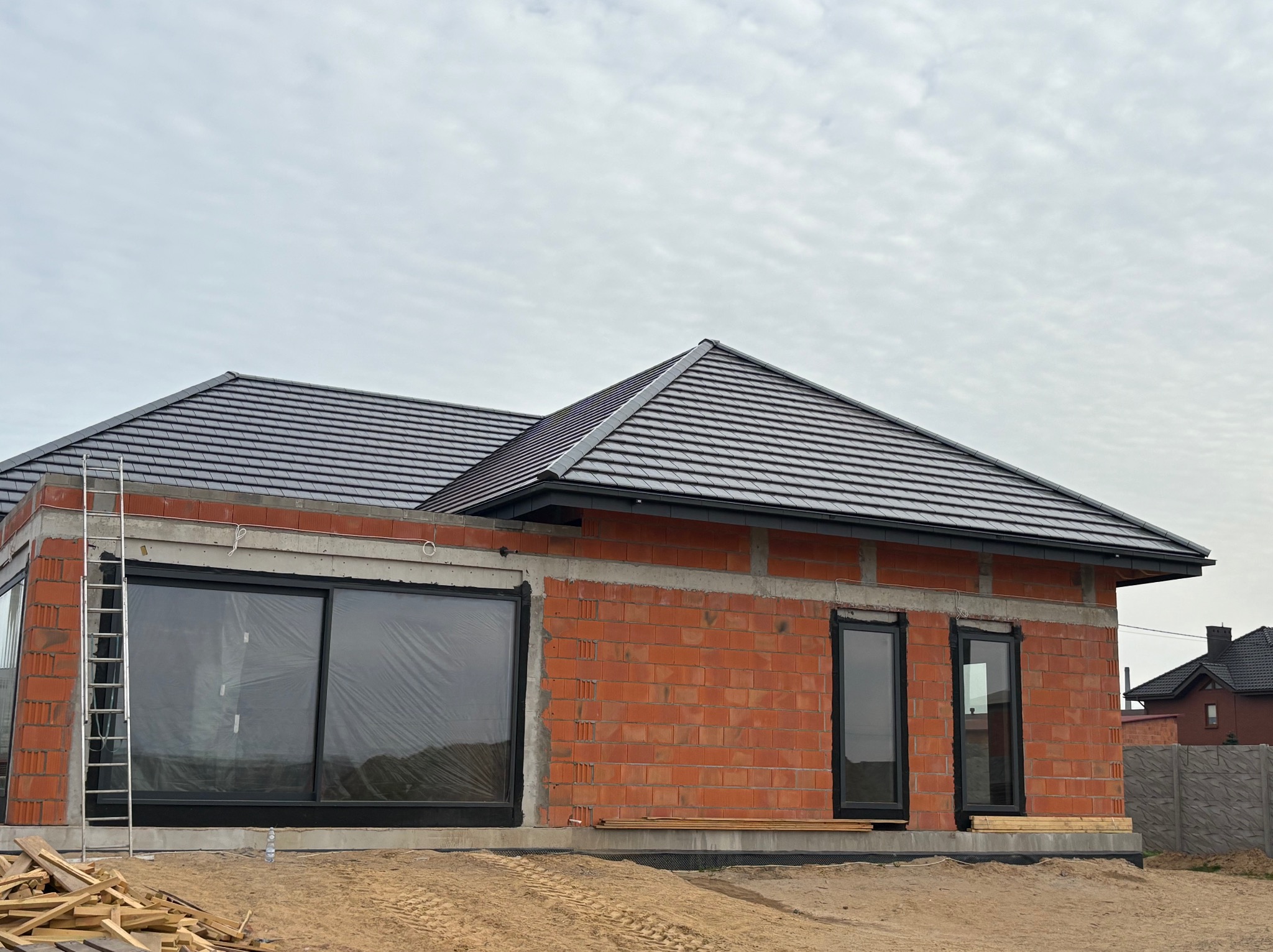 Dom w trakcie budowy z widocznym dachem pokrytym dachówką ceramiczną, oknami zabezpieczonymi folią, czerwoną cegłą i metalową drabiną opartą o ścianę.
