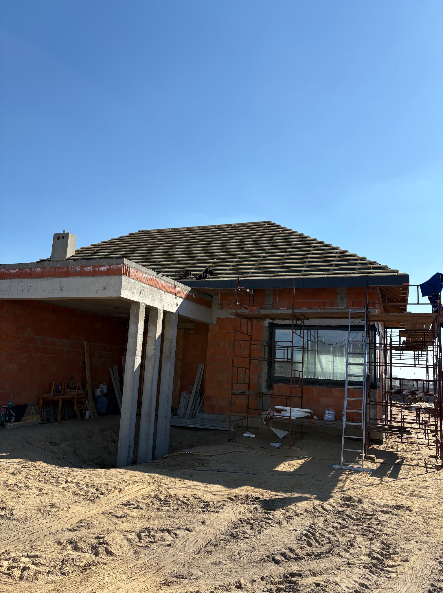 Budowa domu z cegły: widoczny dach z dachówką, rusztowania i elementy konstrukcyjne w słoneczny dzień. Dom w trakcie budowy, zarys okna.