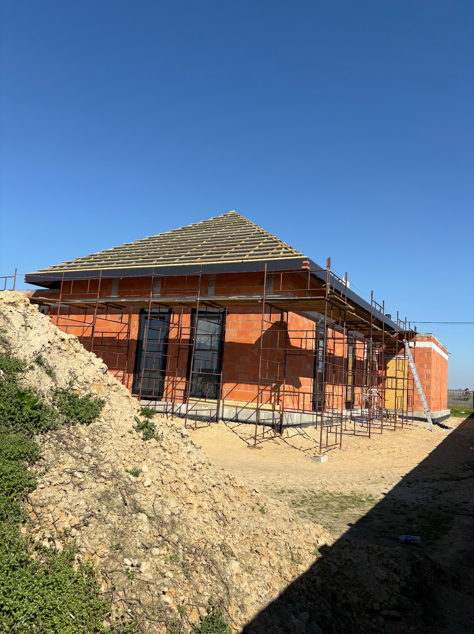 Dom w trakcie budowy: czerwona cegła, czarne okna, rusztowania. Częściowo ułożone pokrycie dachu z deskowaniem. Na pierwszym planie ziemia i zieleń.