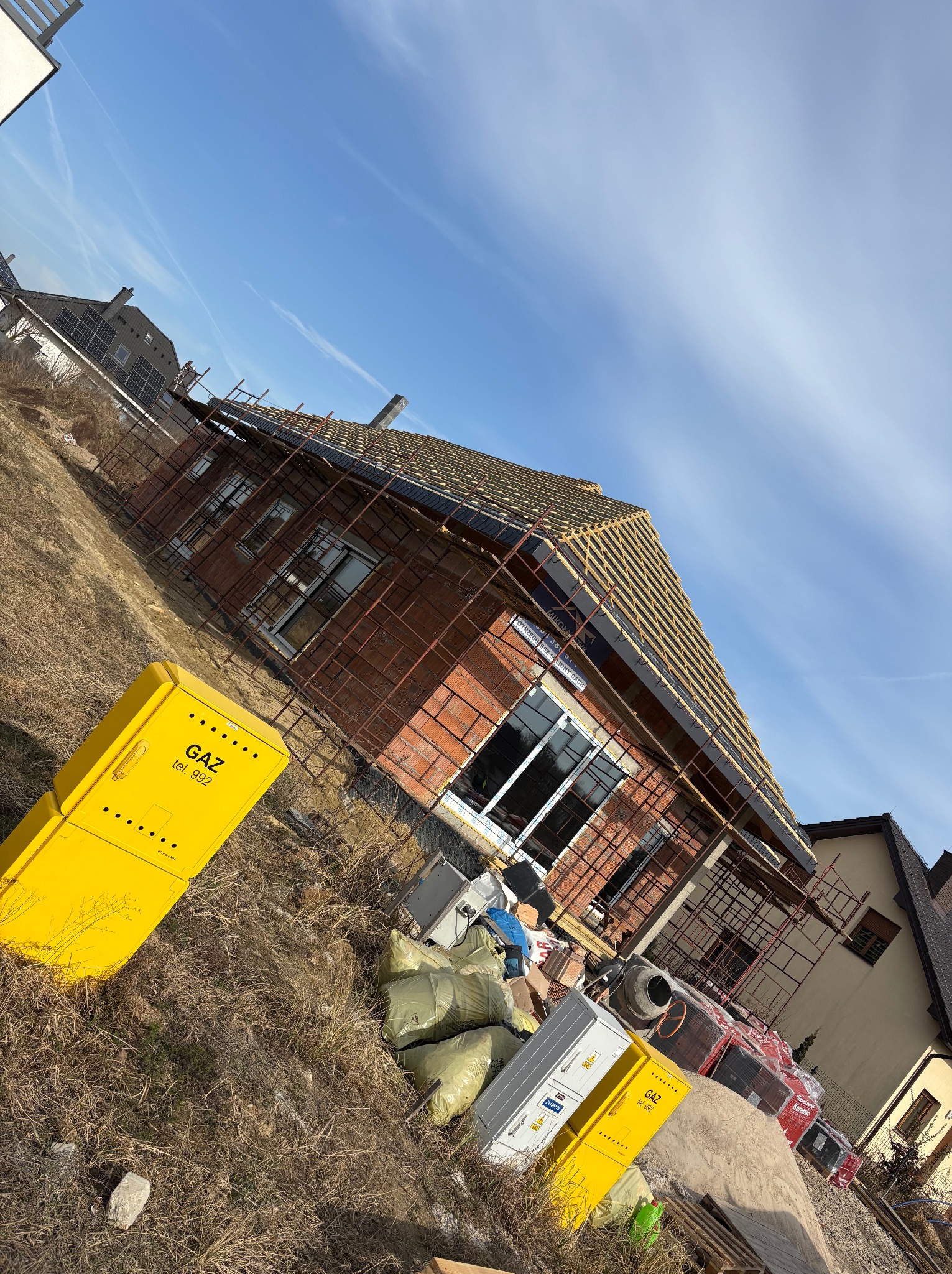 Dom w trakcie budowy z widocznymi rusztowaniami, częściowo ułożonym dachem i skrzynkami gazowymi na pierwszym planie, w słoneczny dzień.