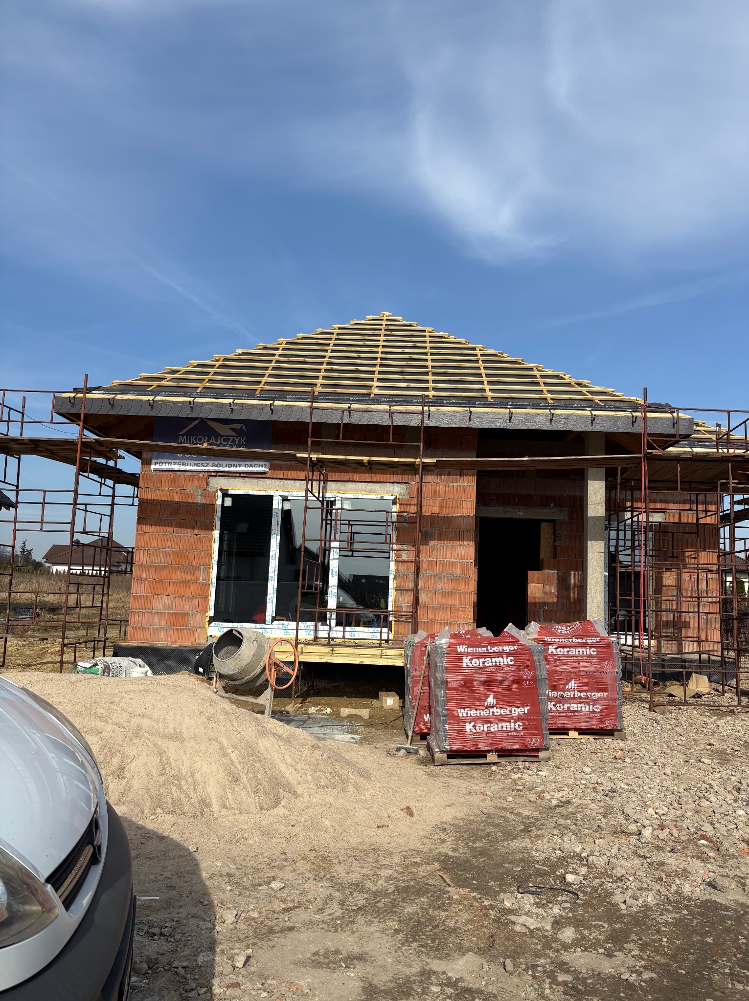 Budowa domu jednorodzinnego w trakcie krycia dachu dachówką ceramiczną Wienerberger Koramic. Widoczne rusztowania, pustaki i betoniarka. Częściowo widoczny samochód.