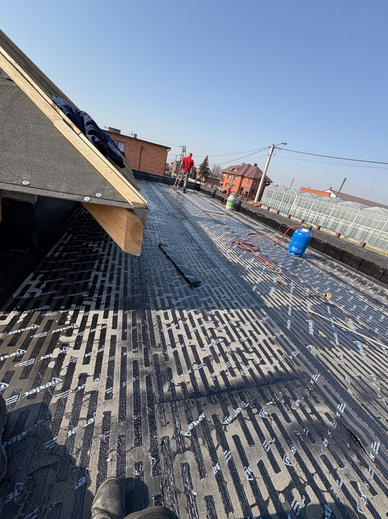 Czarny dach pokryty papą z widocznym wzorem, na którym stoją butle gazowe i narzędzia. W tle domy i szklarnia, a w oddali pracownik w czerwonej koszulce.
