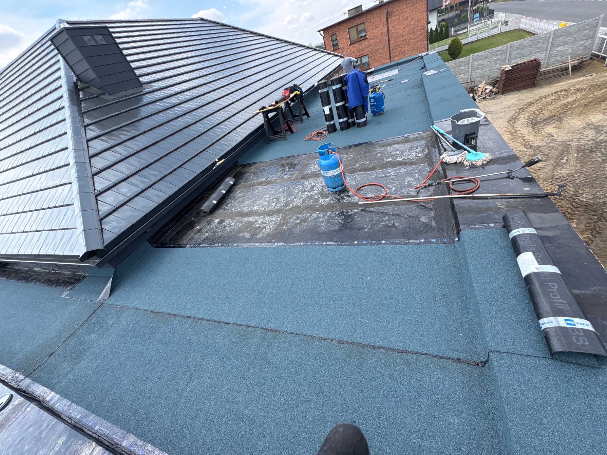 Dach po hydroizolacji papą termozgrzewalną, widoczne butle gazowe, narzędzia i fragment dachu z dachówką ceramiczną. Prace dekarskie w toku.