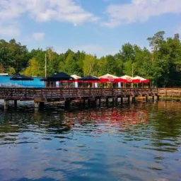 Drewniany pomost nad jeziorem z parasolami, otoczony zielonymi drzewami i błękitnym niebem. W prawym dolnym rogu widoczne logo firmy.